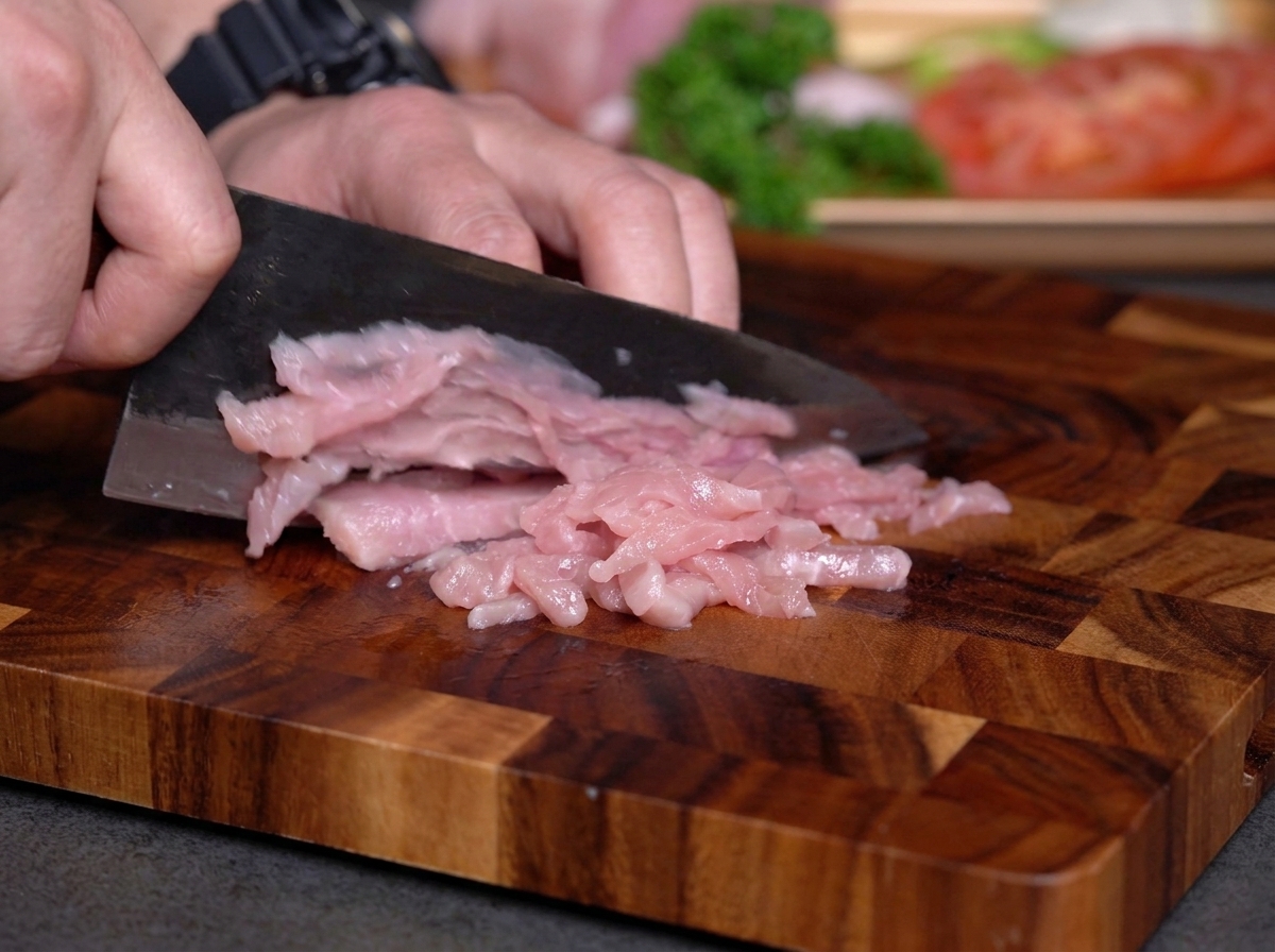 Hands slicing raw pink lean meat into thin strips on a wooden cutting board using a dark metal knife.