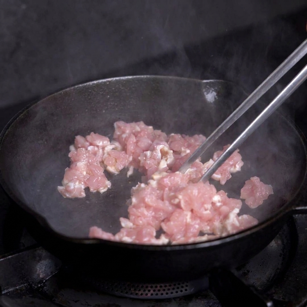 Diced lean meat being cooked in a hot cast iron skillet with metal tongs.