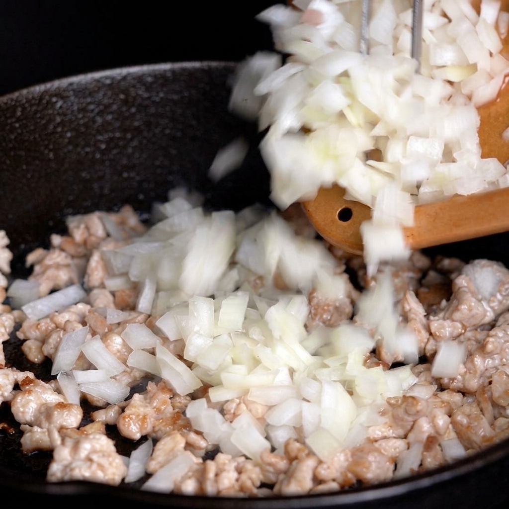 Chopped white onions being added from a cutting board into a skillet containing partially cooked diced meat.