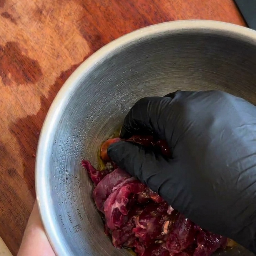 A gloved hand thoroughly mixing raw beef strips with dark marinade in a bowl.