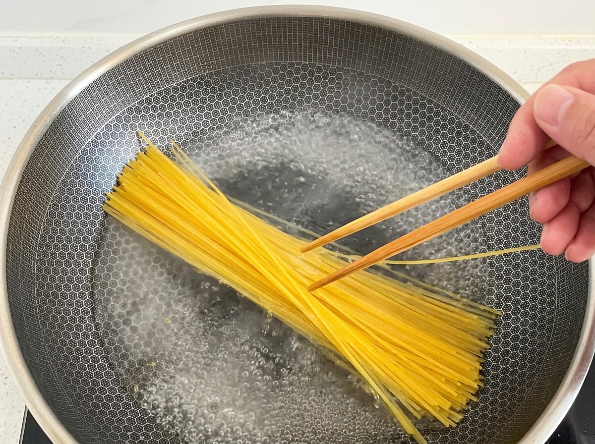 A hand using wooden chopsticks to submerge dry yellow spaghetti noodles into a large wok of vigorously boiling water.