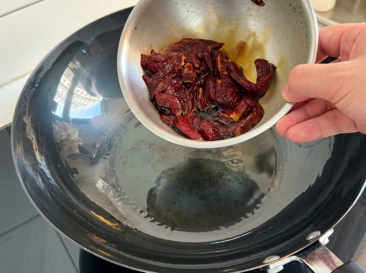 Marinated raw beef strips being poured from a small metal bowl into a hot, oiled wok.
