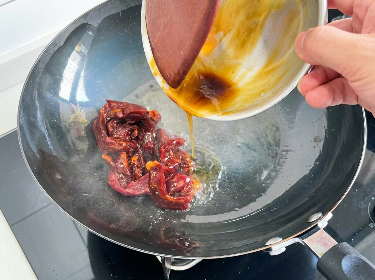Marinated dark red beef strips being poured from a small metal bowl into a hot wok with cooking oil.