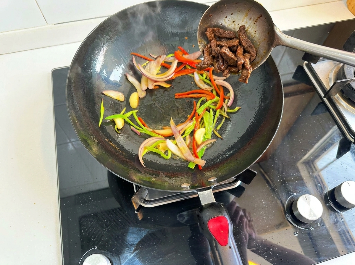 Partially cooked beef strips being added from a metal skimmer back into a wok containing sautéed sliced red and green bell peppers, onions, and garlic.