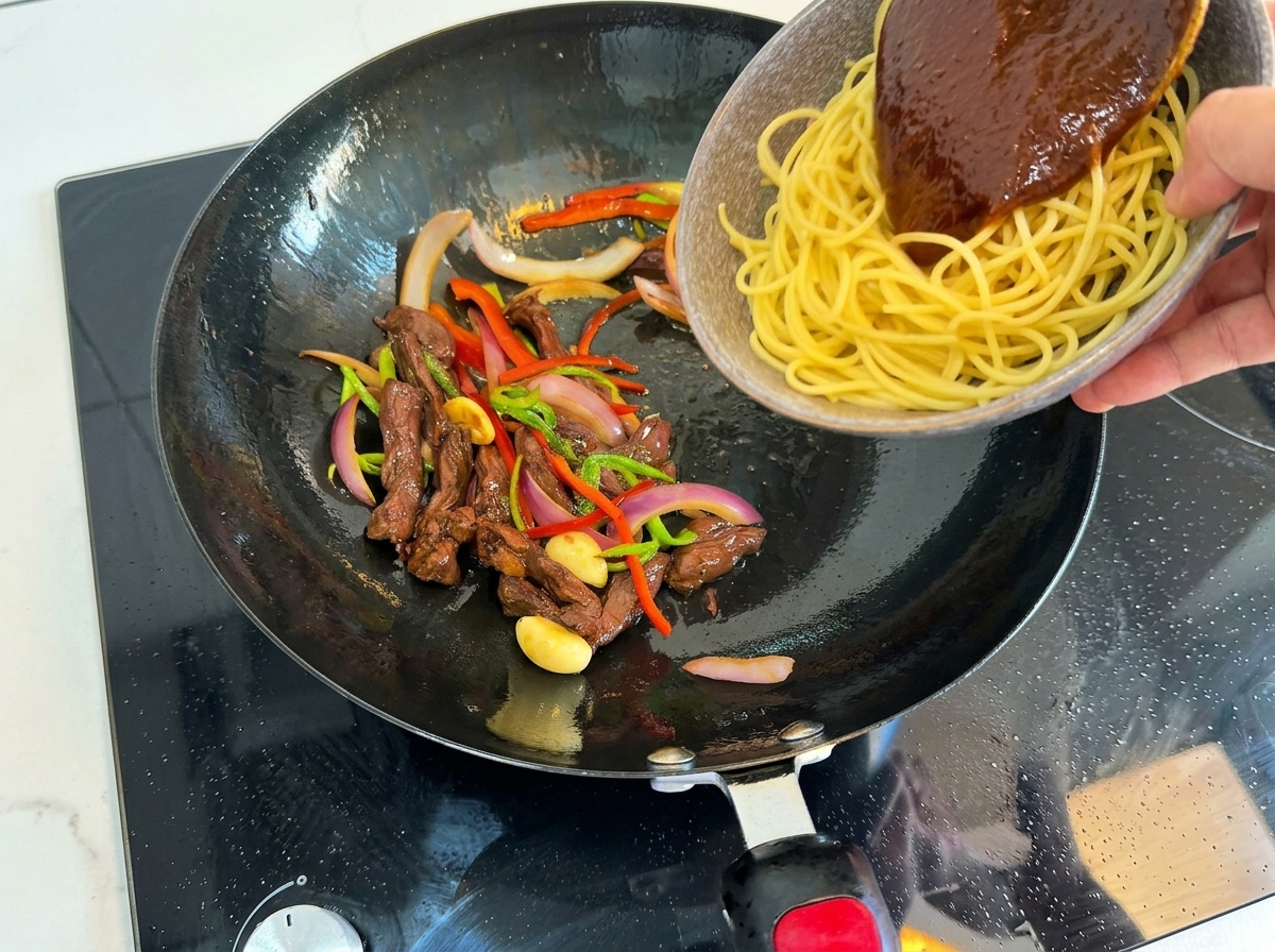 Cooked spaghetti and dark sauce being poured from a bowl into a hot wok containing stir-fried beef, peppers, and onions.