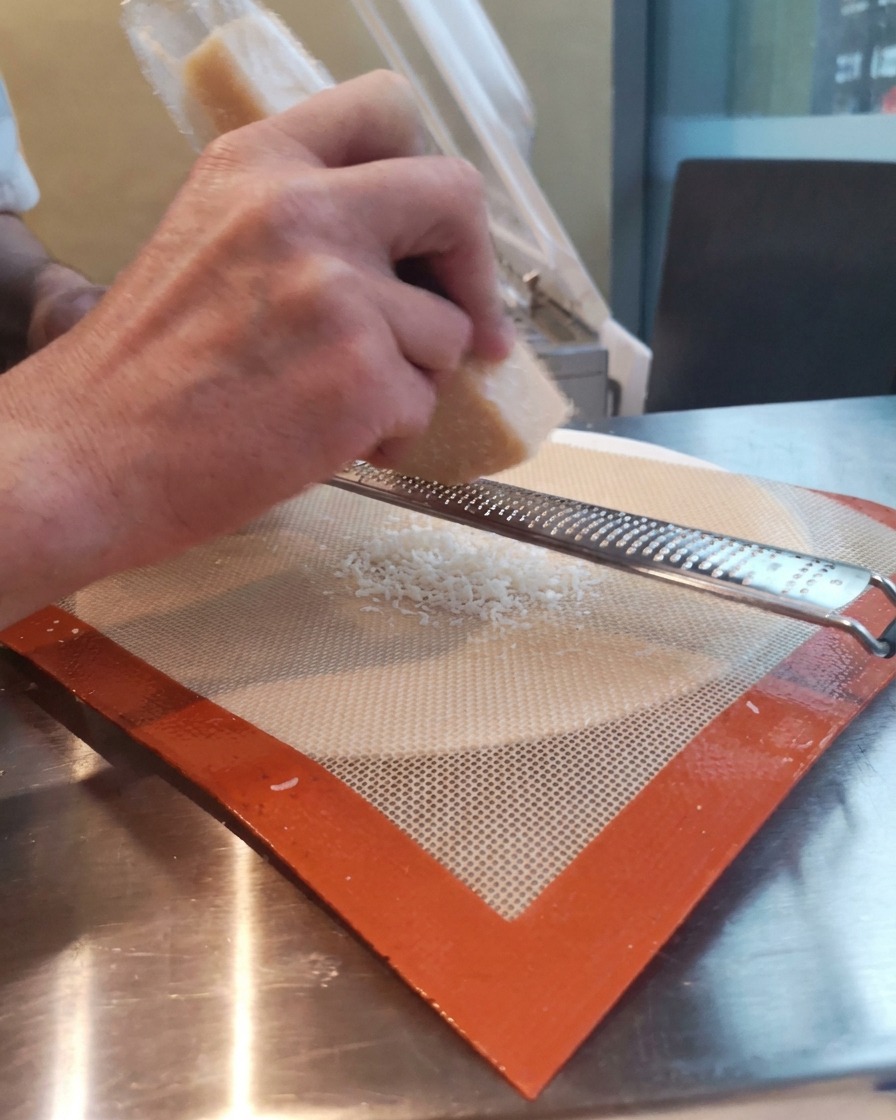 Hands grating a block of Parmesan cheese over a silicone baking mat using a long microplane zester.