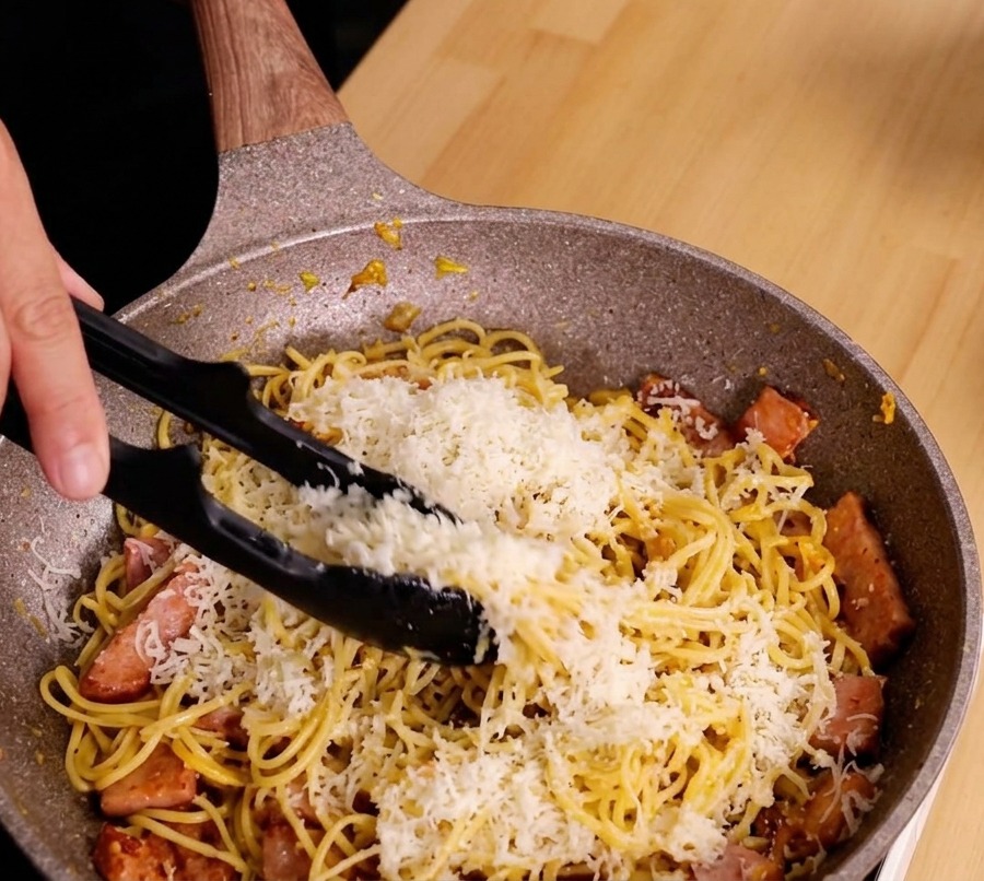 Hands using tongs to actively stir and toss spaghetti, cheese, and bacon in a speckled pan to create a creamy sauce.