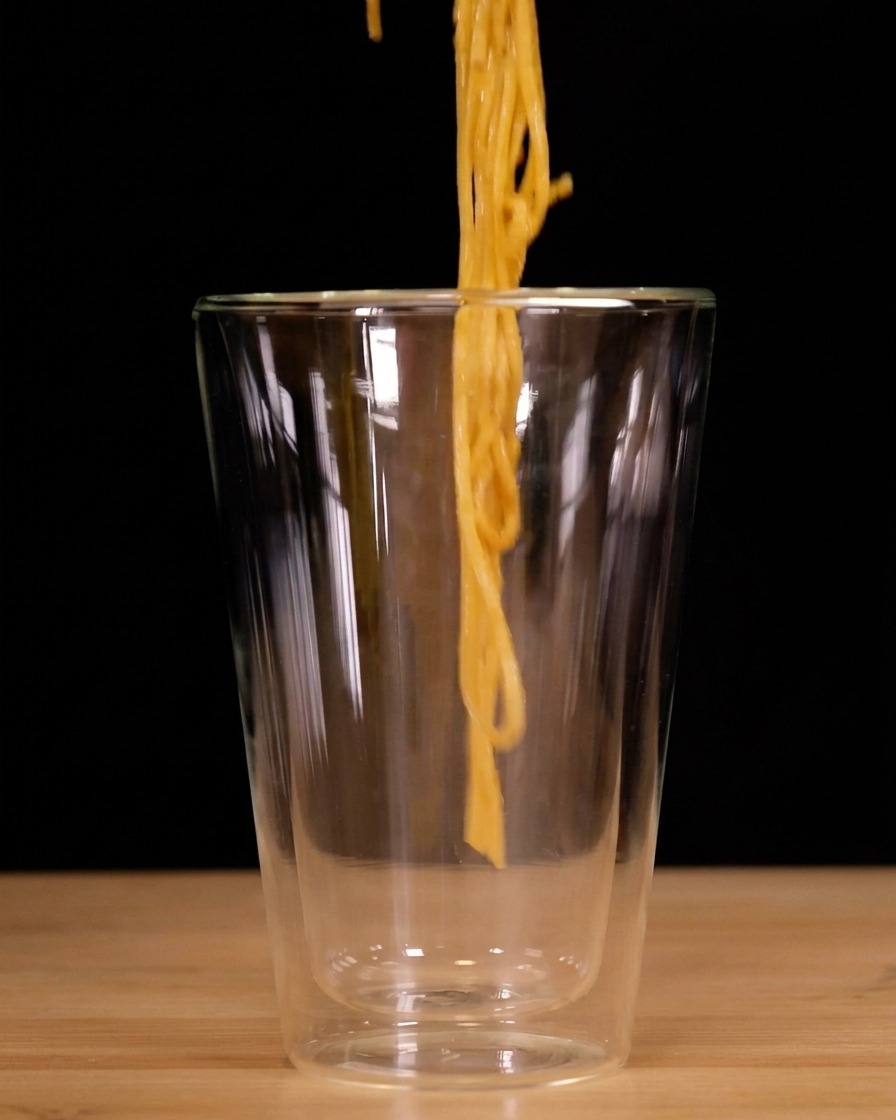 Spaghetti being twirled vertically with tongs and lowered into a tall, clear double-walled glass.