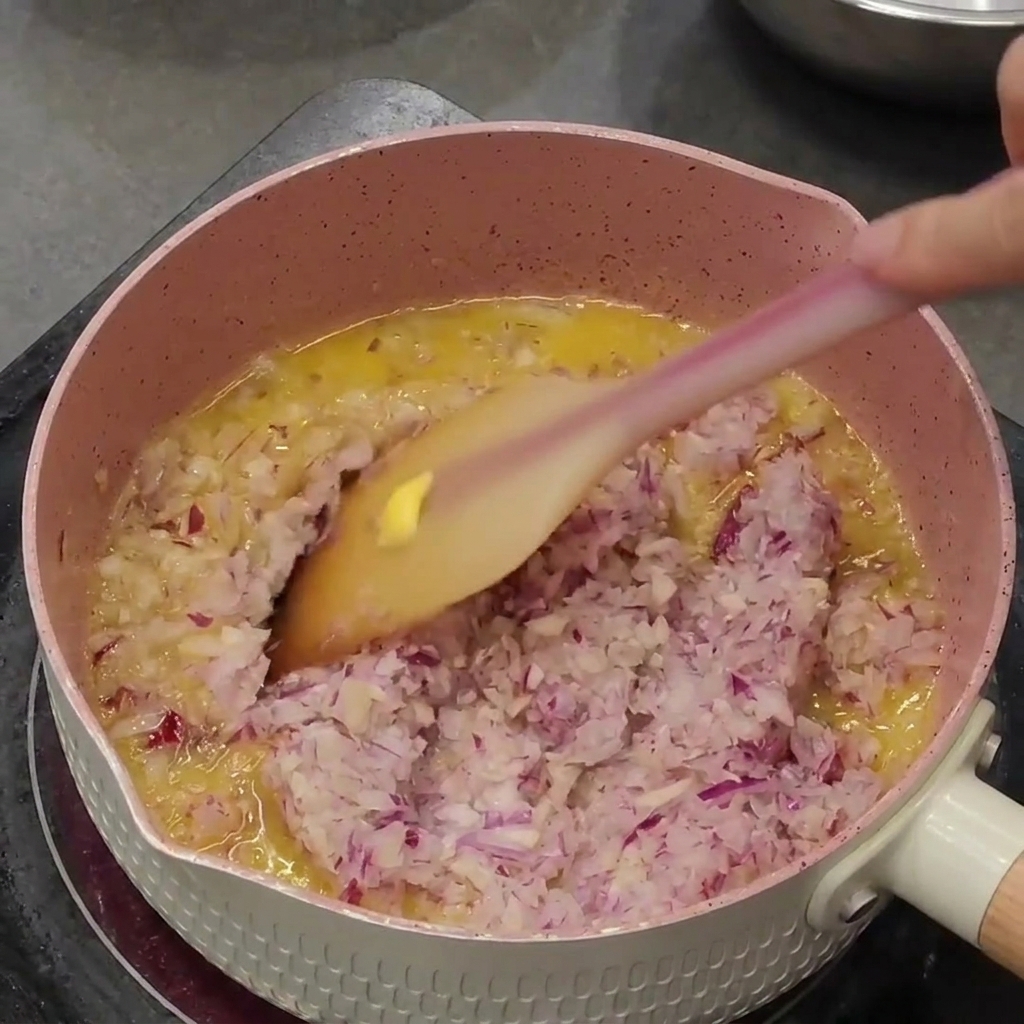 Minced purple onions and garlic being sautéed in melted butter inside a pink saucepan.