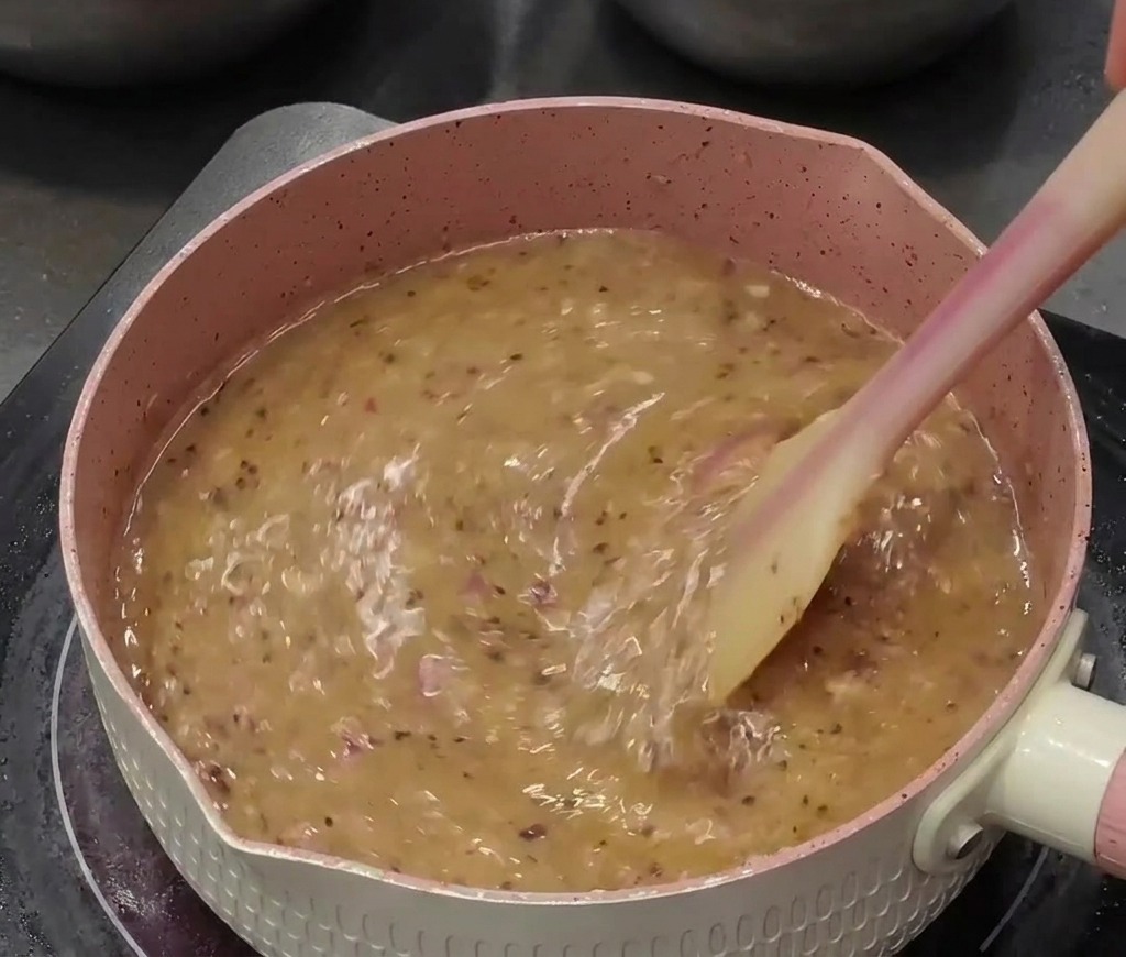 A watery black pepper sauce bubbling in a pink pan, being actively stirred with a silicone spatula.