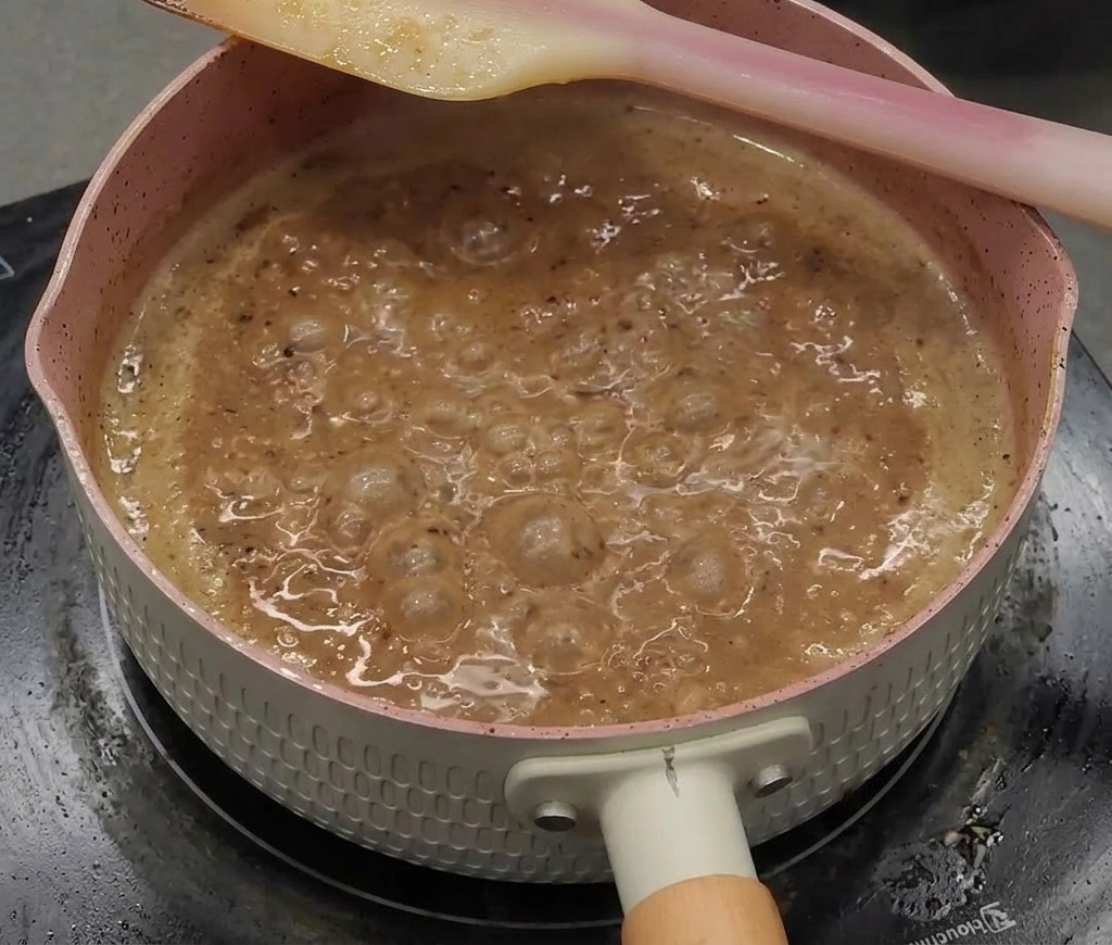 A thick, glossy, dark brown black pepper sauce bubbling vigorously in a pink pan with a spatula resting on the edge.
