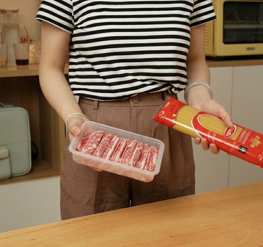 A person holding a package of dry spaghetti and a tray of raw thinly sliced beef.