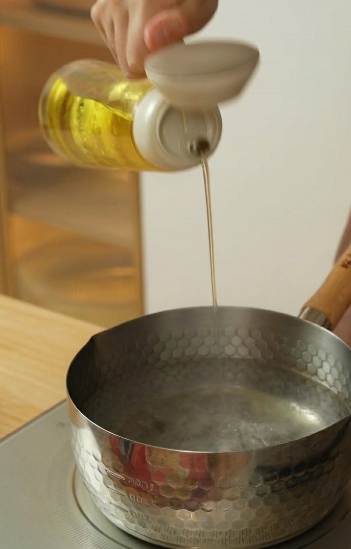 Pouring cooking oil from a bottle into a stainless steel pot of hot water.