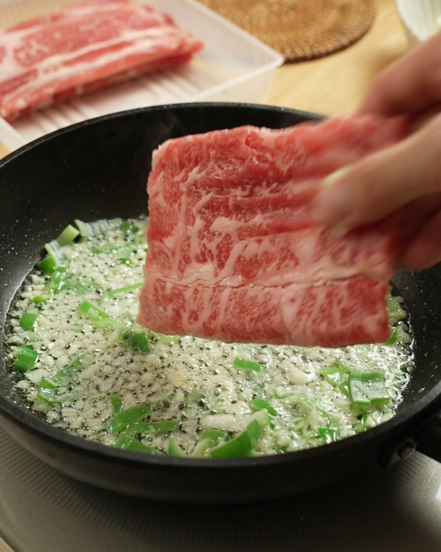 Placing raw, thinly sliced beef into a hot frying pan with sautéed green scallions and garlic.