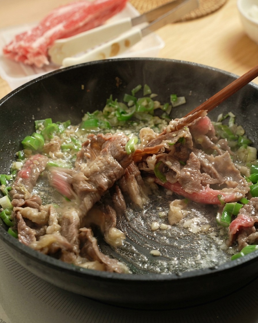 Using chopsticks to stir-fry thin slices of raw beef in a pan with chopped scallions and garlic.
