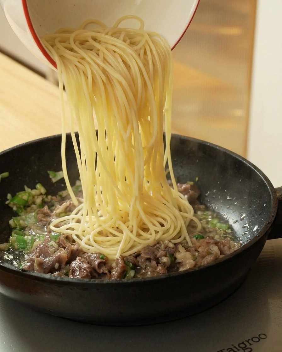 Pouring cooked spaghetti from a bowl into a frying pan containing cooked beef and aromatics.