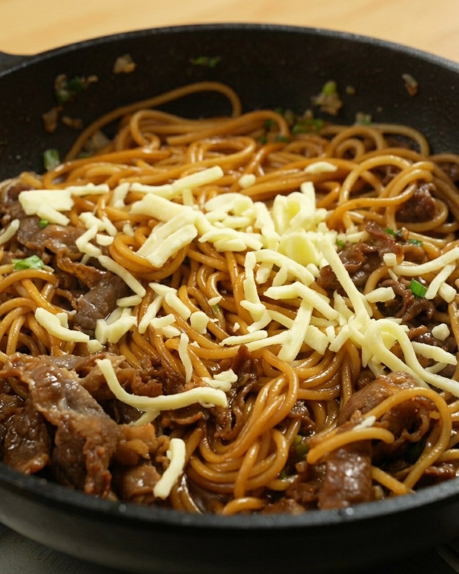 Shredded mozzarella cheese sprinkled over hot stir-fried spaghetti and beef in a pan.