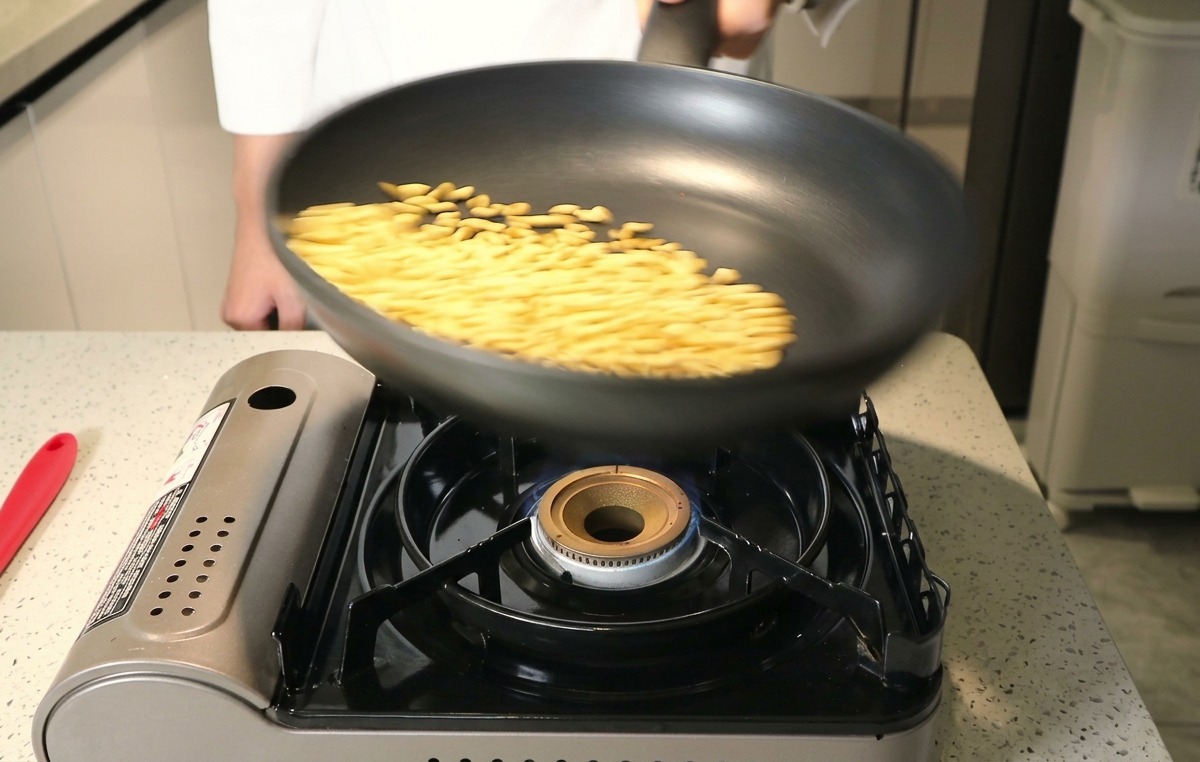 Raw pine nuts being tossed in a black frying pan over a lit portable gas stove.