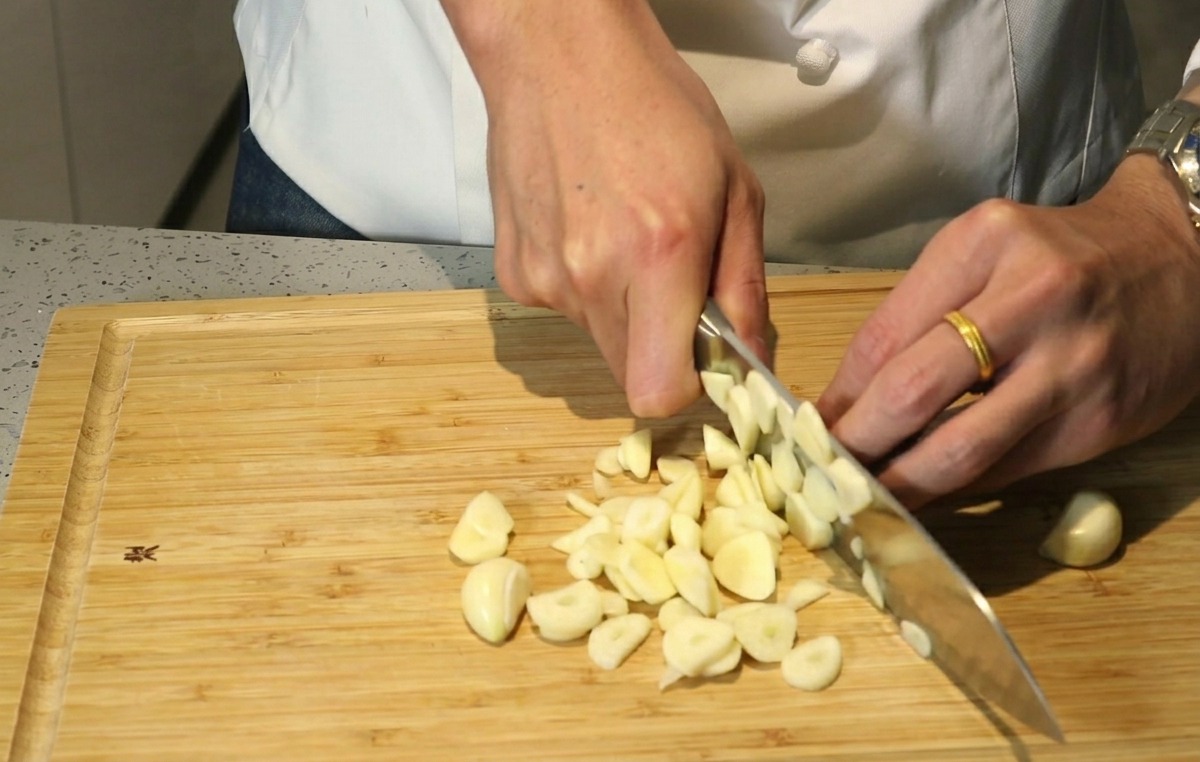 Chefs hands using a knife to roughly chop garlic cloves on a wooden cutting board.