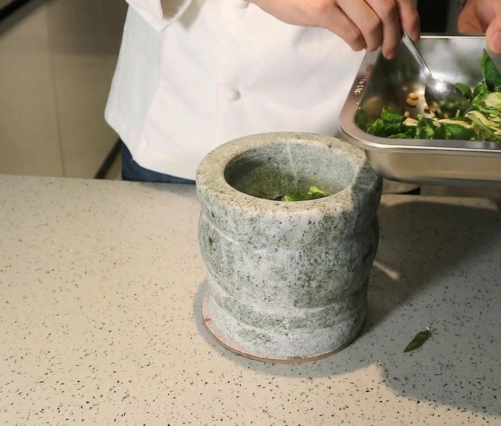 Chef scraping a mixture of fresh basil, pine nuts, and garlic from a metal tray into a grey stone mortar.