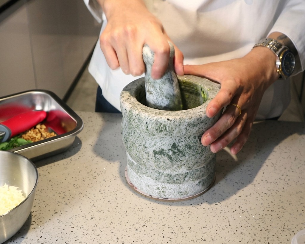 A person using a stone mortar and pestle to grind fresh ingredients into a green pesto paste.