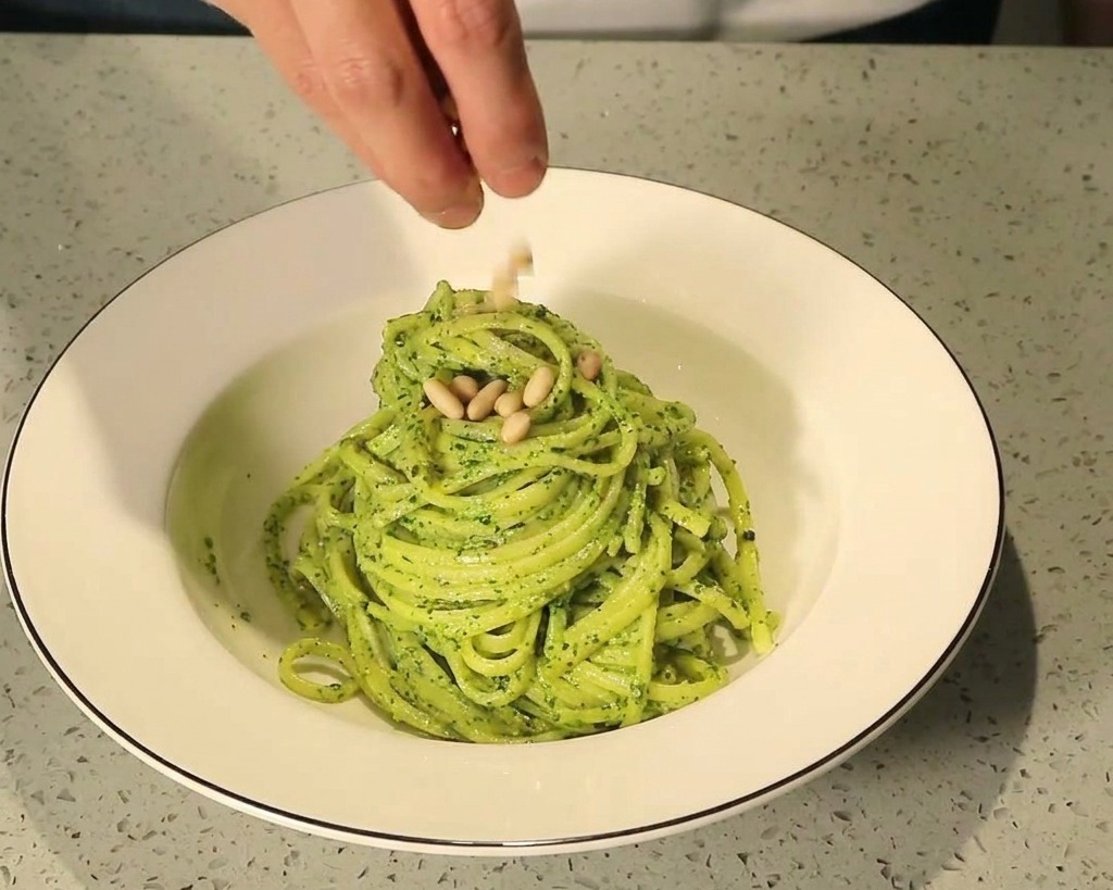 Fingers sprinkling toasted pine nuts over a neatly twirled mound of green pesto pasta in a white ceramic bowl.