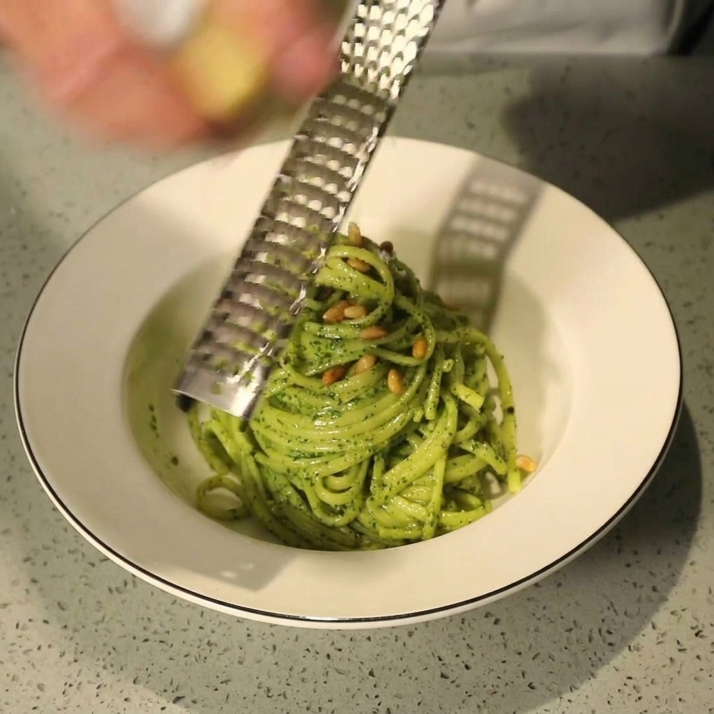 A hand using a metal grater to shave fresh Parmesan cheese over a plated bowl of green pesto pasta sprinkled with pine nuts.