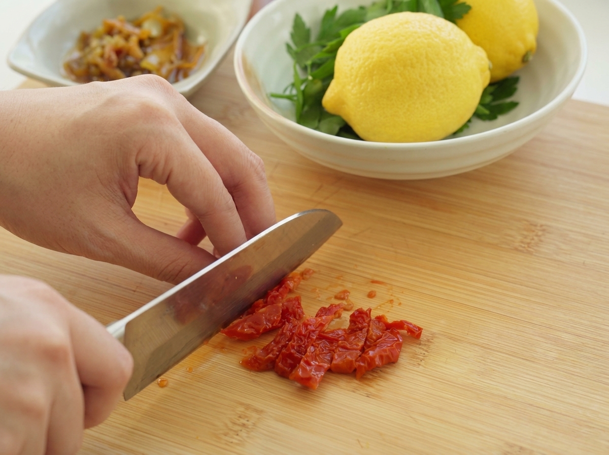 A person using a chefs knife to finely chop vibrant red sun-dried tomatoes on a wooden cutting board.