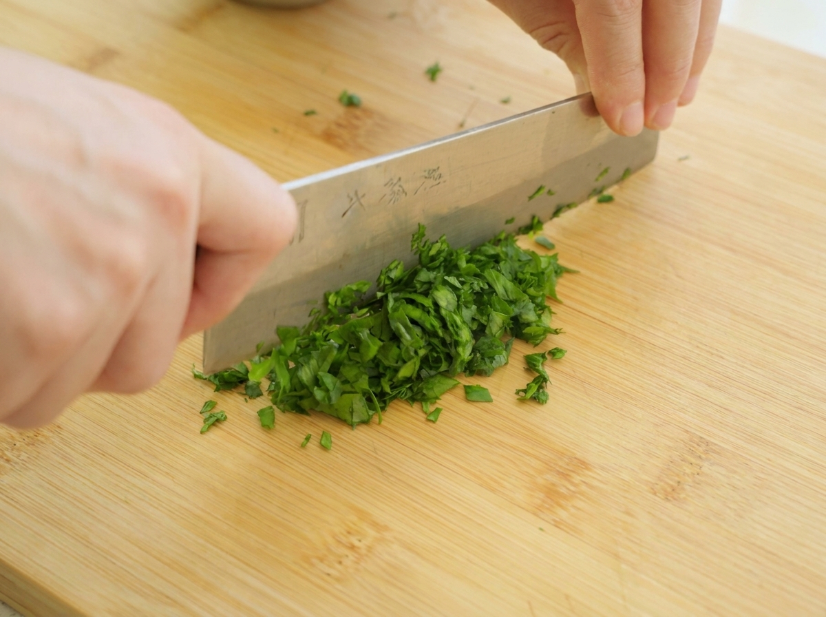 Hands holding a large kitchen knife, finely chopping fresh green parsley leaves on a wooden cutting board.