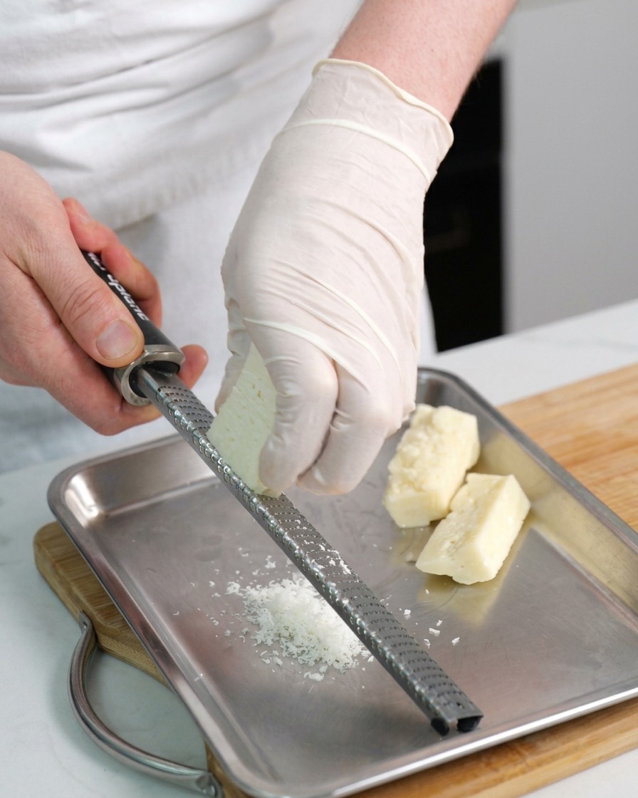 Gloved hands using a handheld microplane grater to shred a block of Pecorino cheese onto a metal tray.