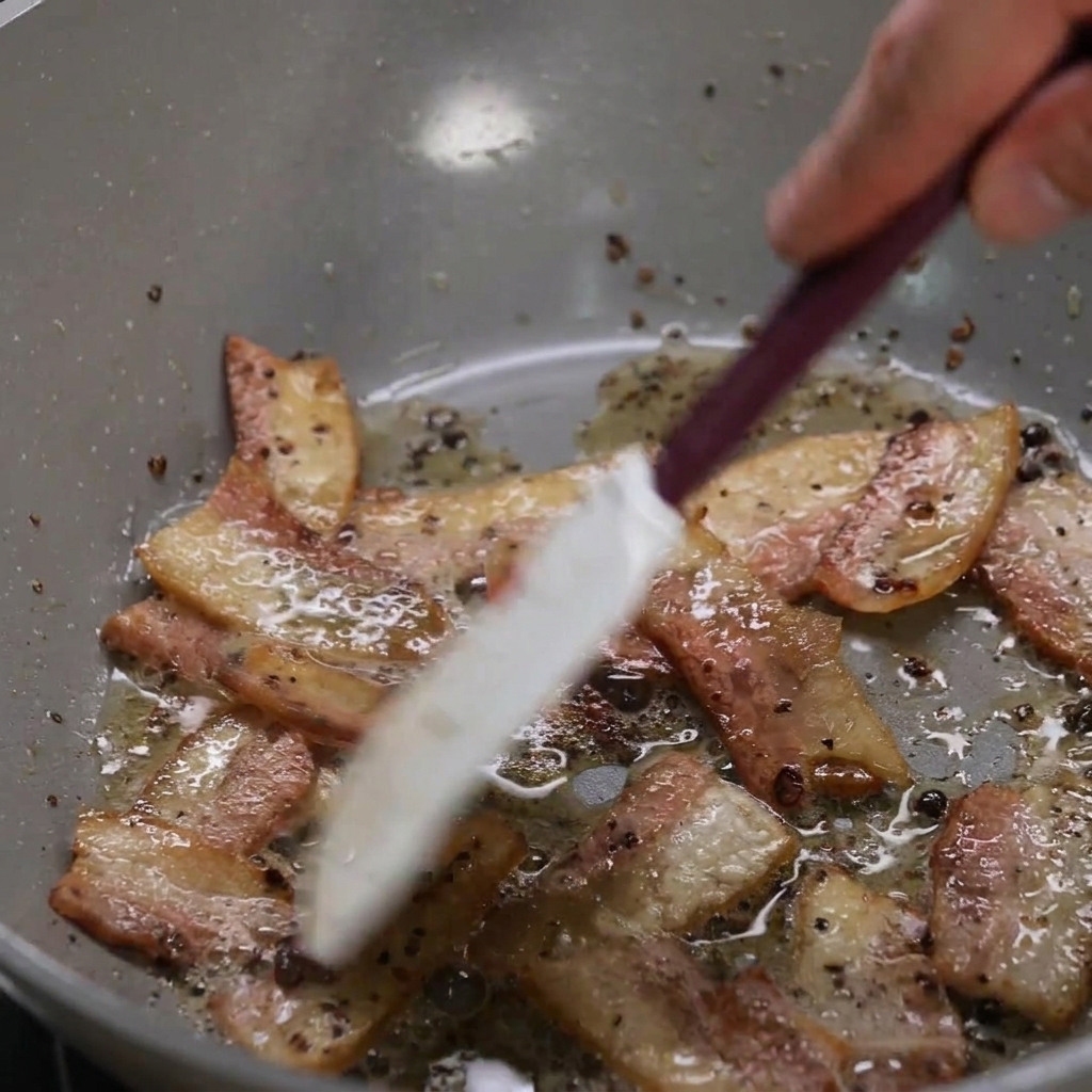 Sliced guanciale browning in a pan with its fat rendering out, being stirred with a light-colored spatula.
