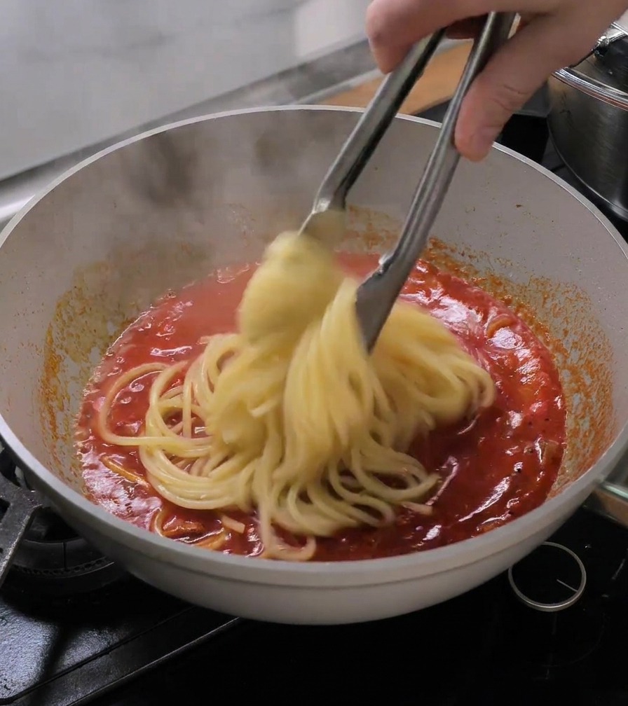 Metal tongs transferring cooked bucatini pasta directly into a pan of simmering red tomato sauce.