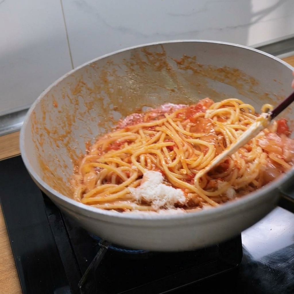 Tossing bucatini pasta with rich red tomato sauce and melting cheese in a white pan.