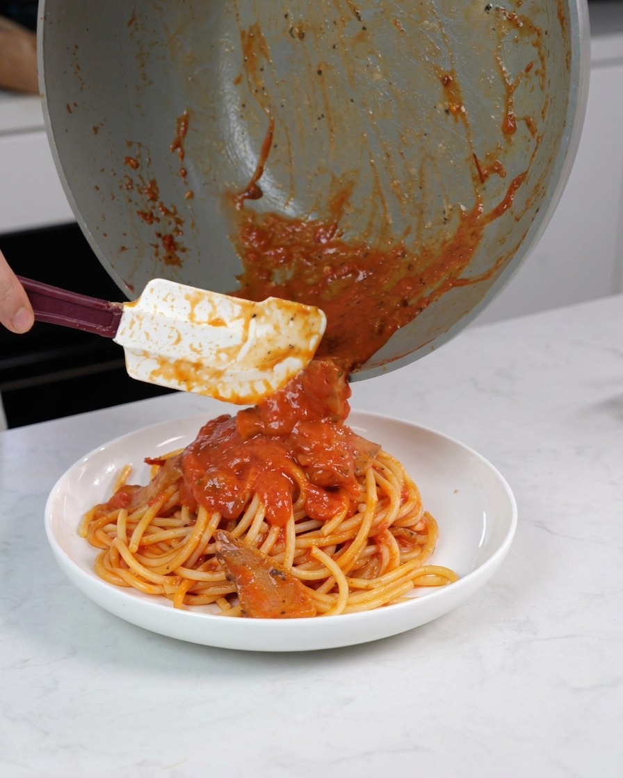 A hand using a white spatula to scrape thick red tomato sauce with pieces of meat from a pan onto a serving of cooked bucatini pasta in a white bowl.