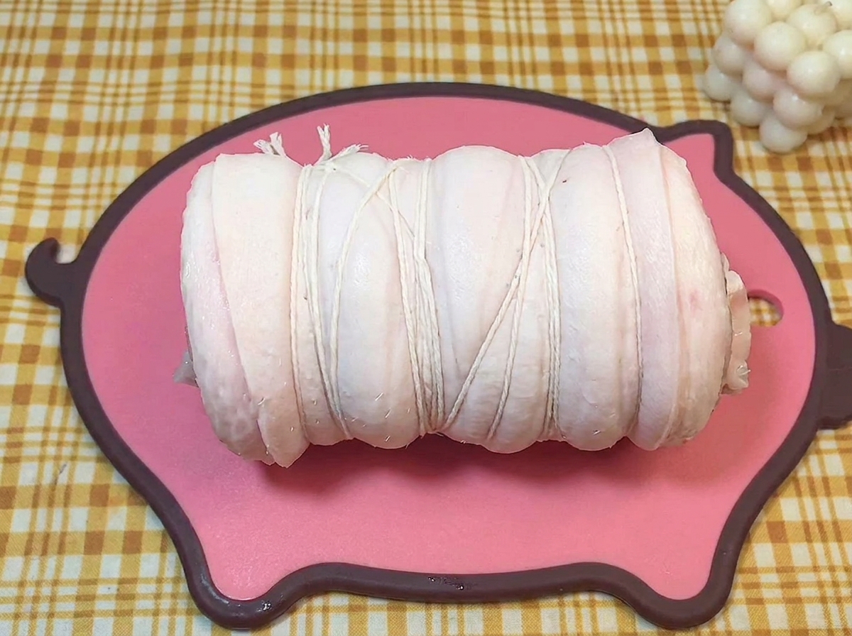 A cylinder of raw pork belly firmly bound with white butchers twine resting on a pink board.