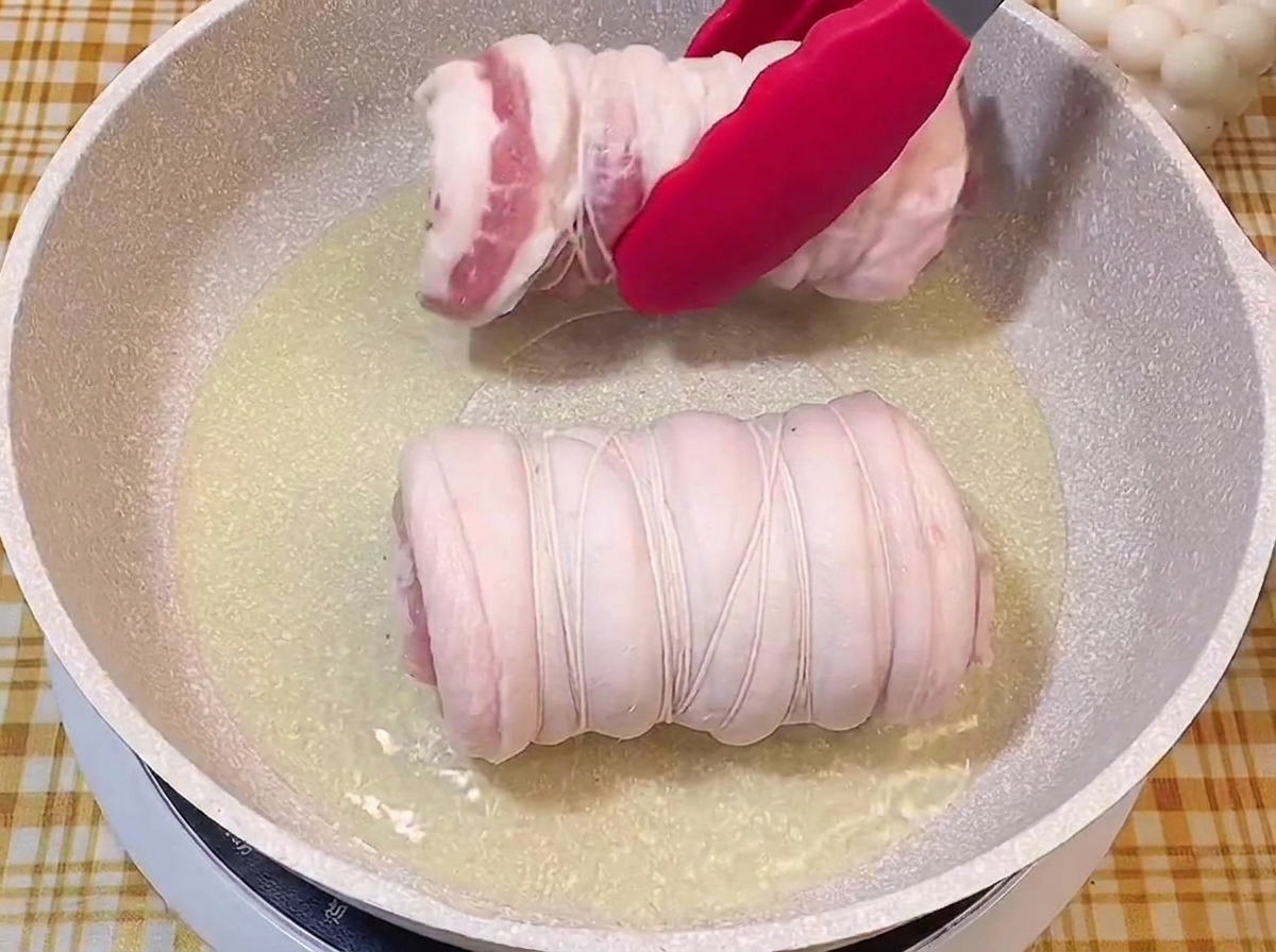 Red tongs placing a tied raw pork roll into a sizzling pan of oil next to another roll.