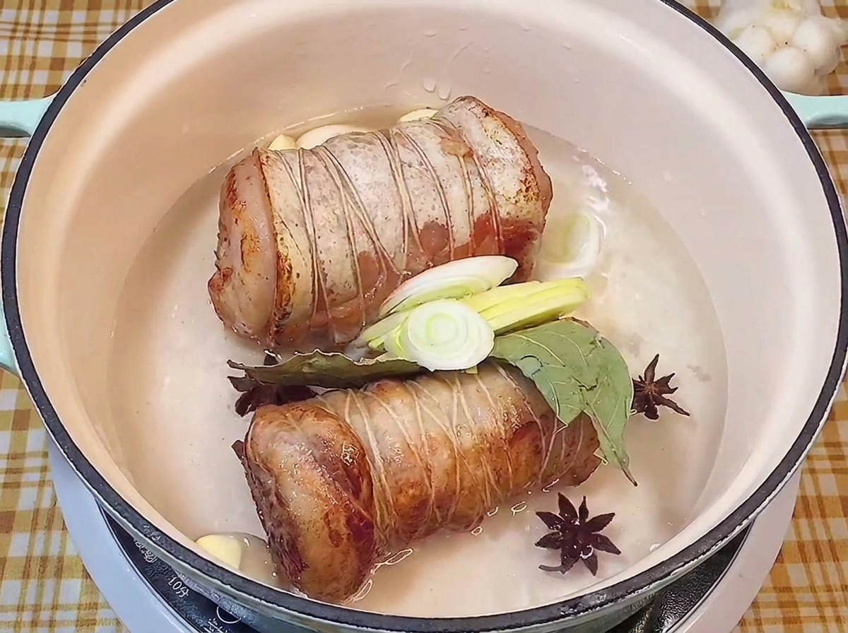 Browned pork rolls resting in a pot of liquid with star anise, bay leaf, garlic, and fresh leeks.