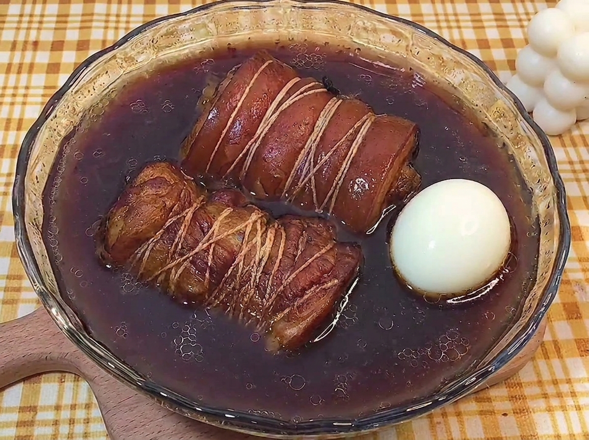 Two rolled braised pork bellies and a whole boiled egg resting in a shallow glass bowl of dark braising liquid.