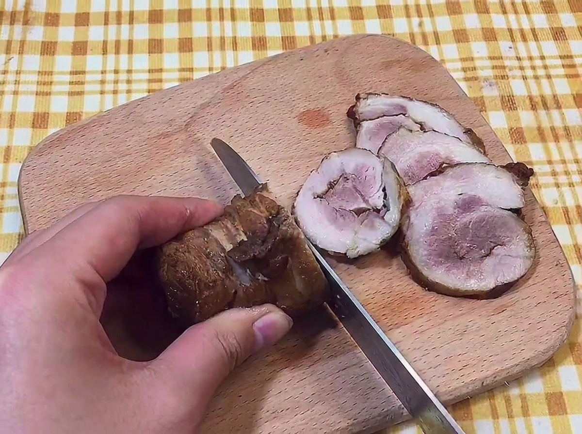 Hand holding a knife to slice a roll of braised pork belly into thin rounds on a wooden cutting board.