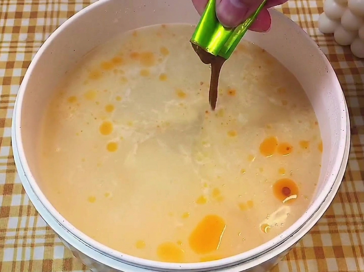 Hand squeezing a packet of dark, concentrated liquid seasoning base into a pot of simmering, light-colored broth.