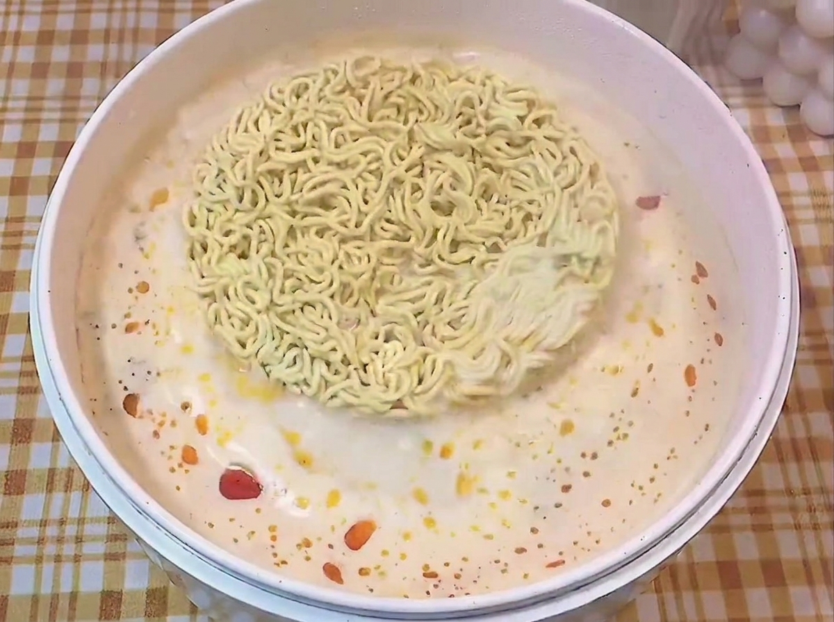 A round block of dry instant ramen noodles floating in a white pot filled with simmering, seasoned broth.