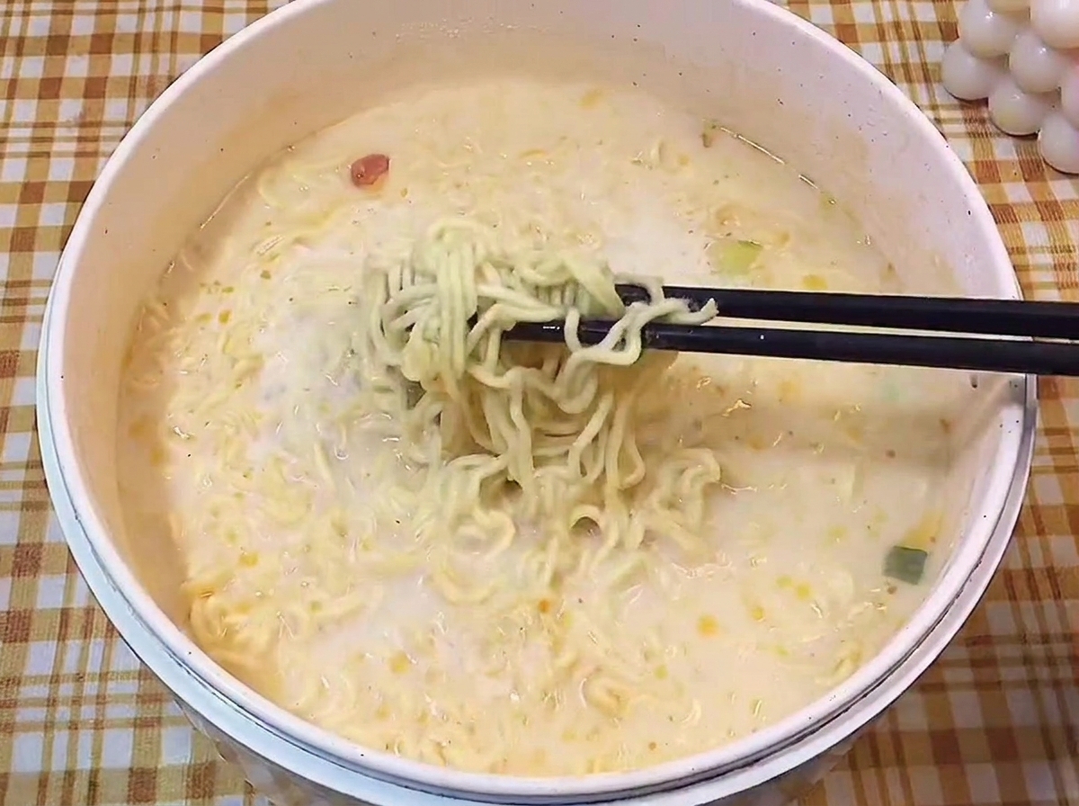 Black chopsticks lifting a portion of cooked, wavy ramen noodles out of a milky, creamy broth.