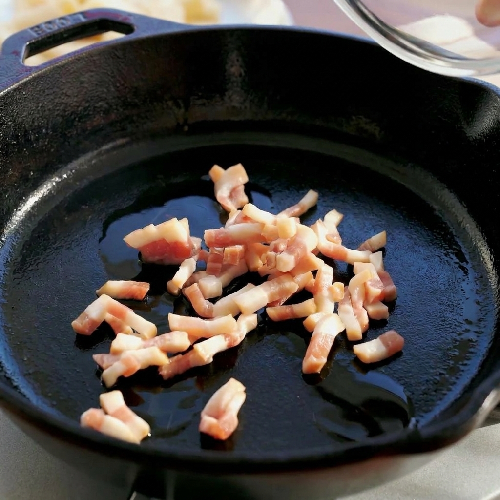 Diced raw guanciale being poured from a glass bowl into a dark cast iron skillet.