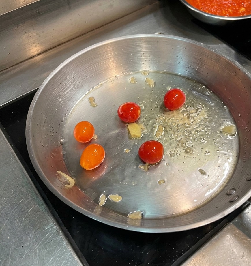 Whole cherry tomatoes blistering in hot oil in a wide stainless steel pan.