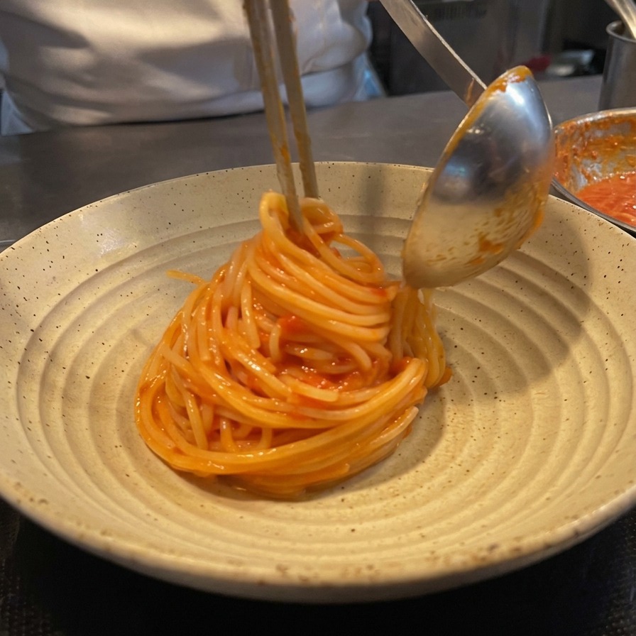 A chef using tongs and a metal ladle to twirl tomato spaghetti into a neat mound on a rustic ceramic plate.