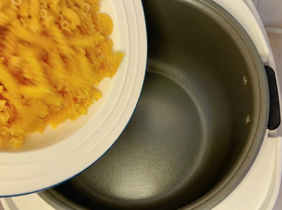 Dry fusilli pasta being poured from a white plate into a dark, empty rice cooker pot.
