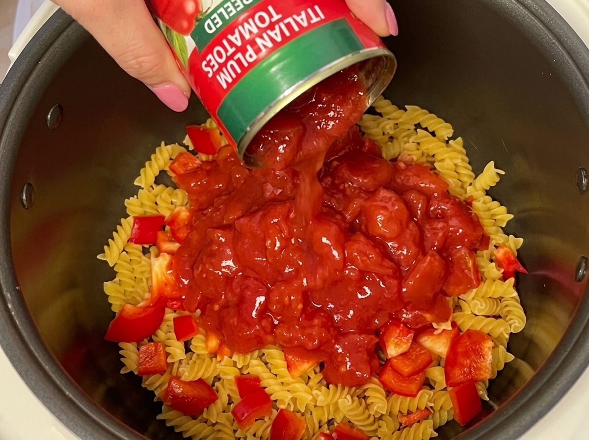 A hand pouring a can of Italian plum peeled tomatoes over dry fusilli pasta and diced red bell peppers.
