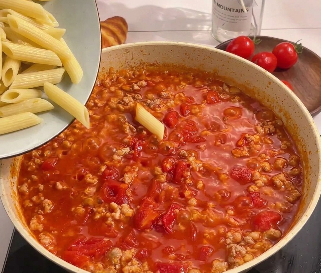 Boiled penne pasta being poured from a bowl into a simmering pan of tomato and meat sauce.