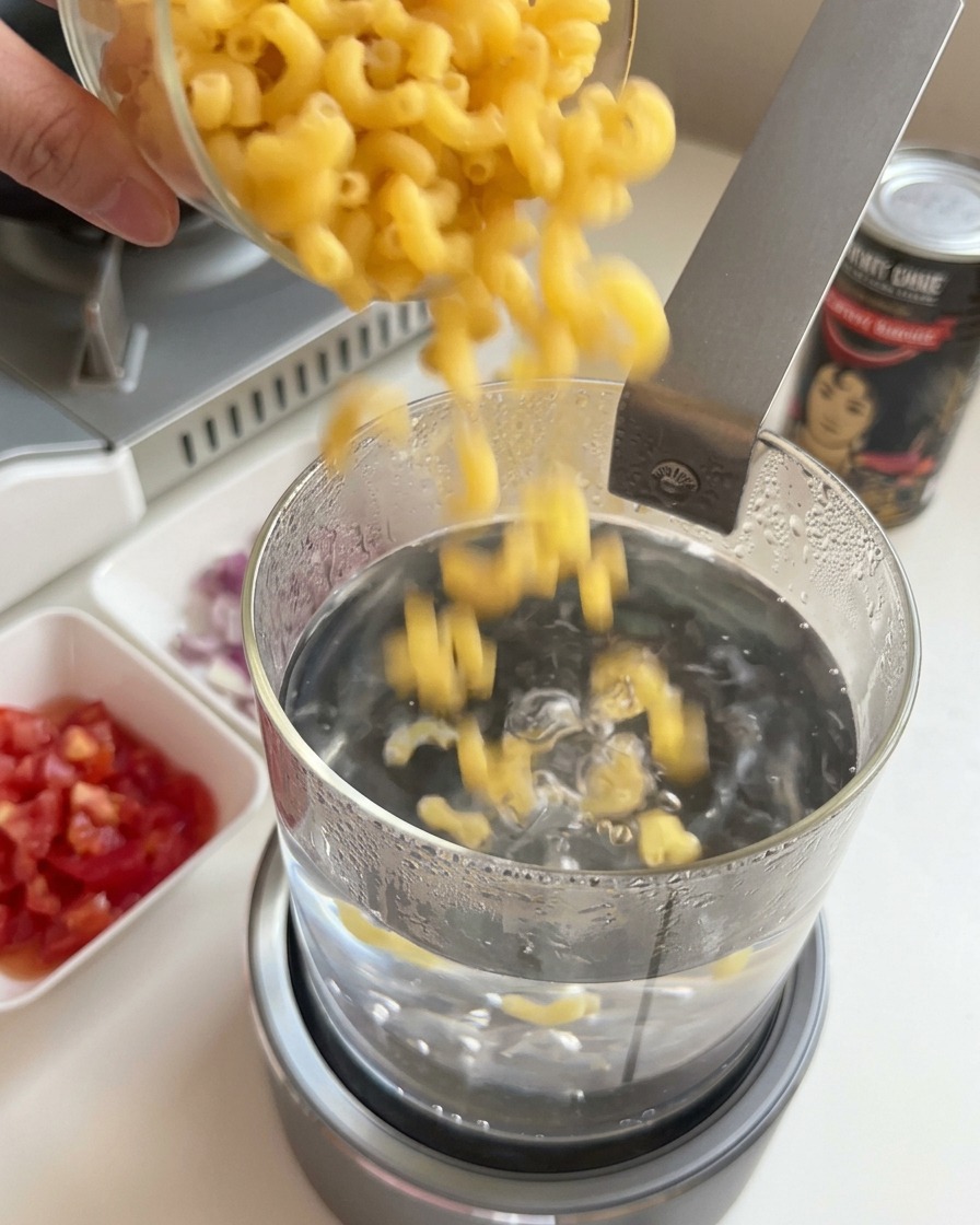 Pouring dry elbow macaroni into a glass pot of boiling water.