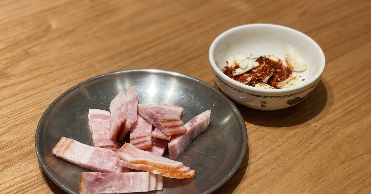 Sliced raw bacon on a metal plate next to a small bowl of sliced garlic and chili flakes resting on a wooden table.